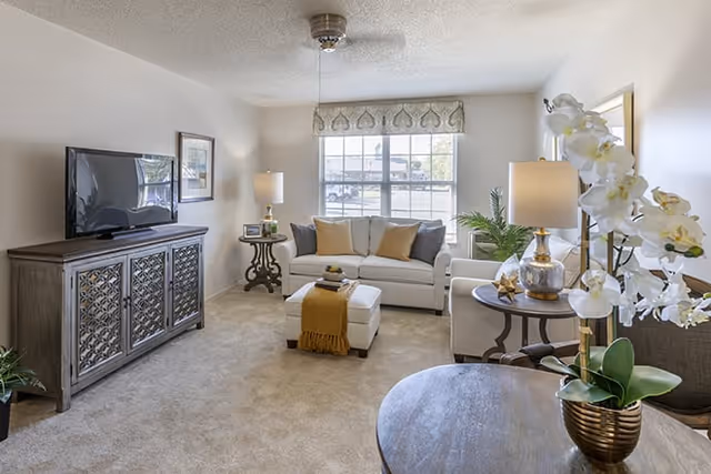 A bright and cozy living room with a large window letting in natural light. The room features a white sofa with multiple cushions, a matching ottoman with a yellow throw, a wooden TV cabinet with a flat-screen TV, two side tables with lamps, a round wooden table with a potted orchid plant, and a beige carpeted floor.