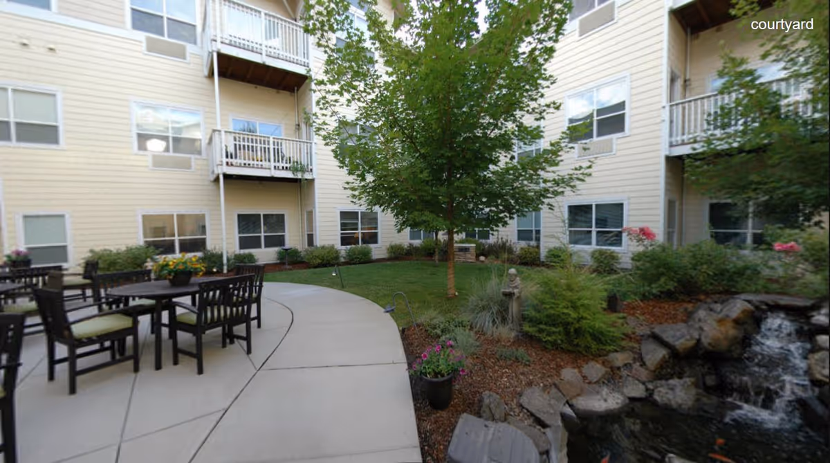 Courtyard with patio tables and chairs, a central tree, landscaped beds and a small pond beside a multi-story residential building.