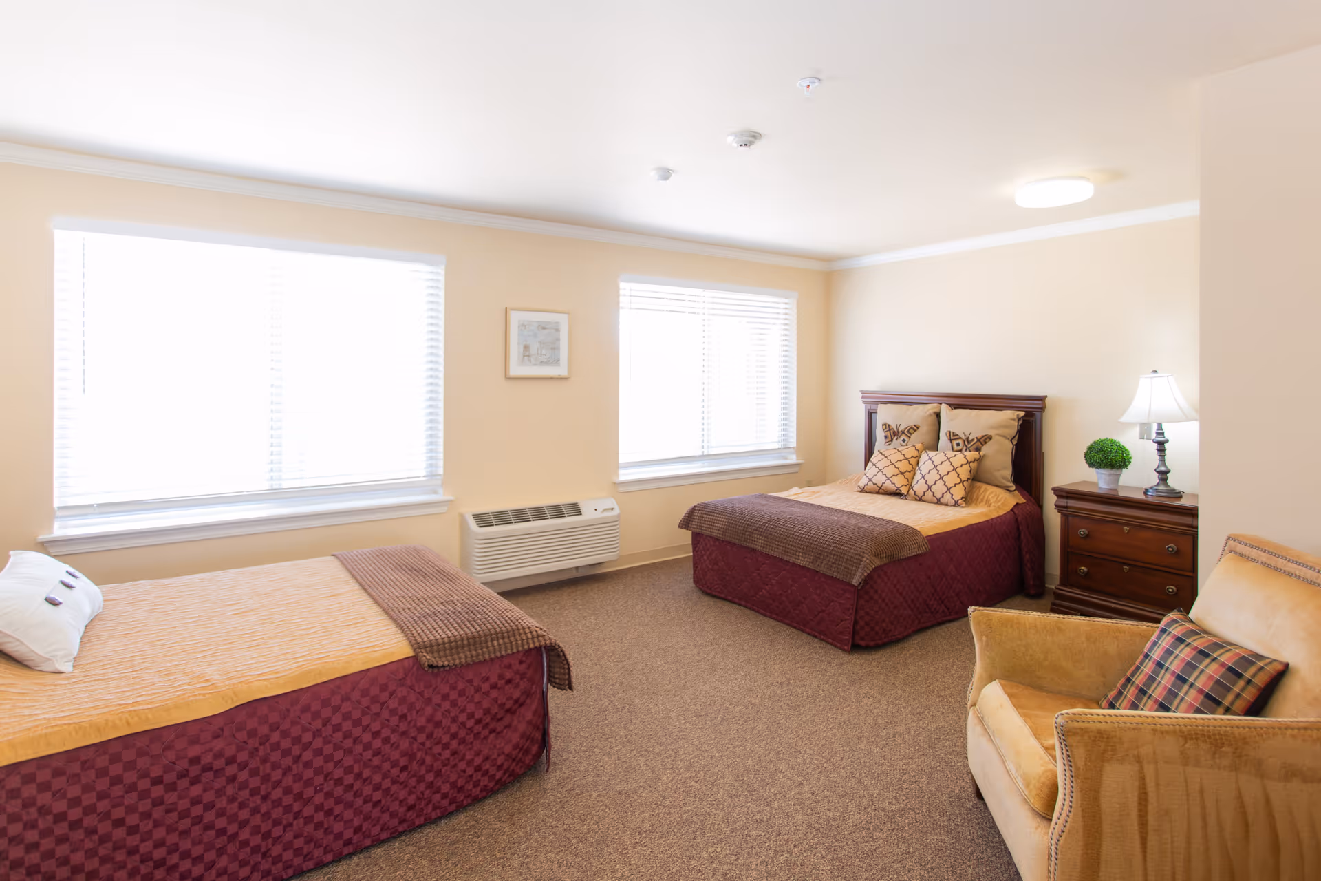 A bright and clean bedroom with two beds, each covered with a maroon and beige bedspread. There is a wooden nightstand with a lamp and a small green plant between the beds. A comfortable armchair with a plaid pillow is positioned in the corner. Two large windows with white blinds allow natural light to fill the room.