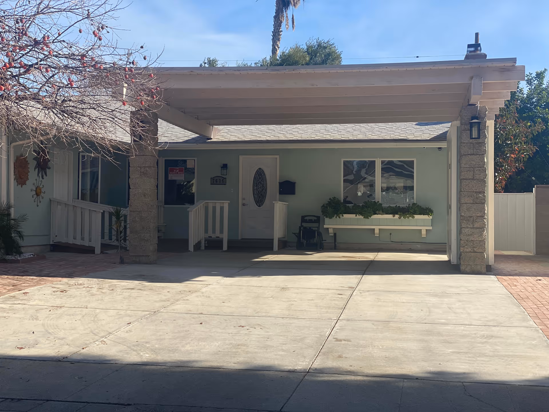 Front exterior view of a single-story assisted living facility with a covered driveway, a white door with an oval glass panel, a window with a planter box beneath it, and a wheelchair parked near the entrance. There are stone pillars supporting the roof of the covered area and some trees and plants around the building.
