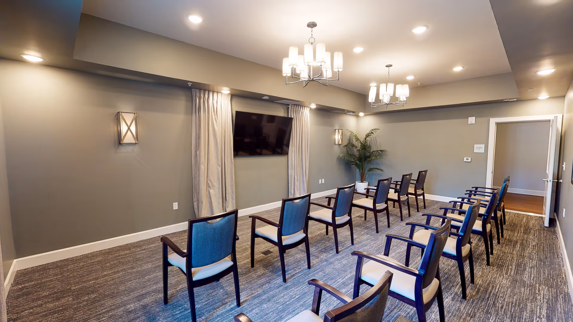 A conference or meeting room with two rows of wooden chairs with light cushions facing a wall-mounted flat screen TV. The room has gray walls, carpeted floor, two chandeliers, wall sconces, and a potted plant in the corner. There is an open door leading to another room.
