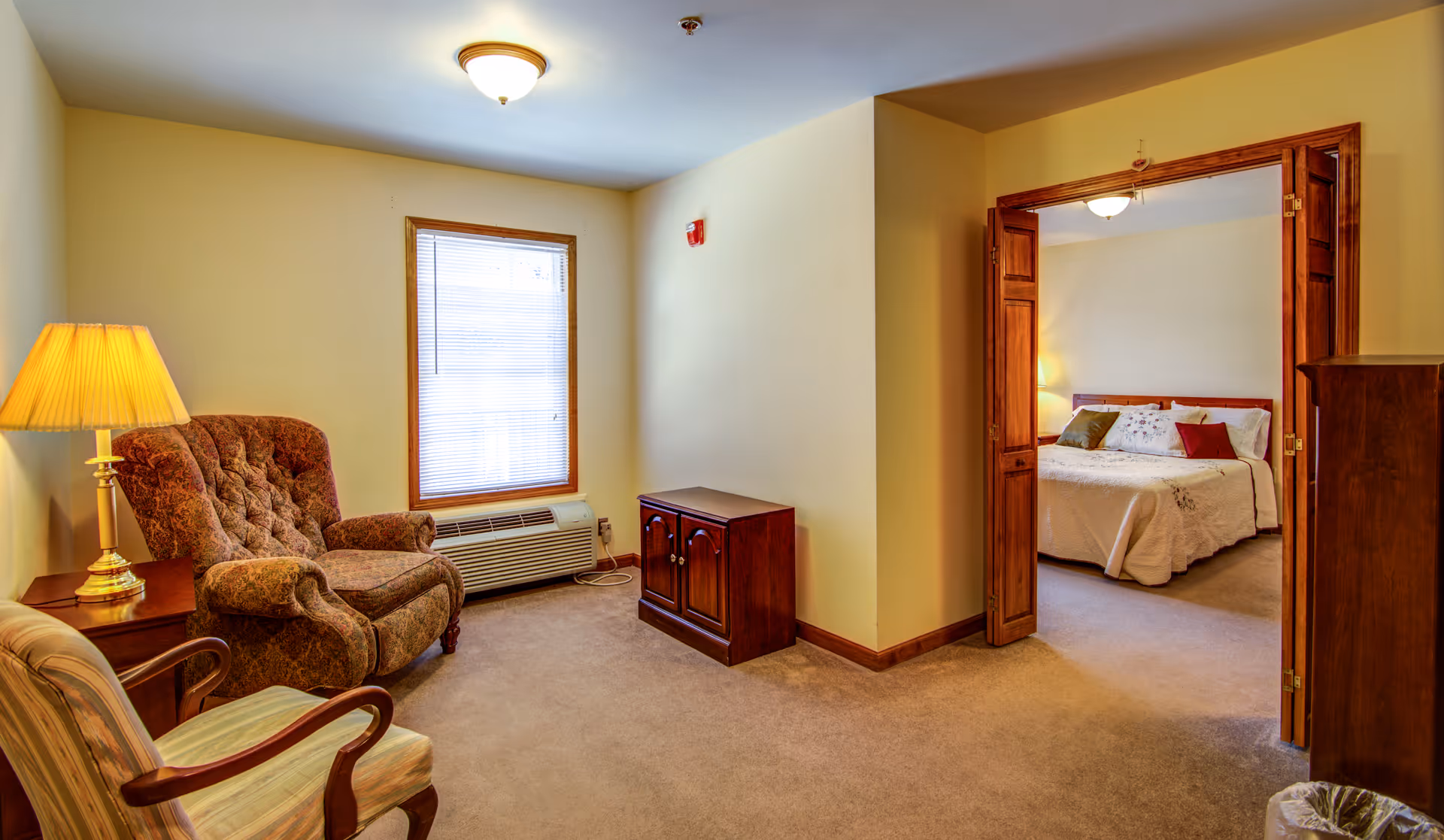 A cozy sitting area with two armchairs and a side table with a lamp, next to a window with blinds. The room has beige walls and carpeted floor. Through an open wooden double door, a bedroom with a bed, pillows, and a nightstand with a lamp is visible.