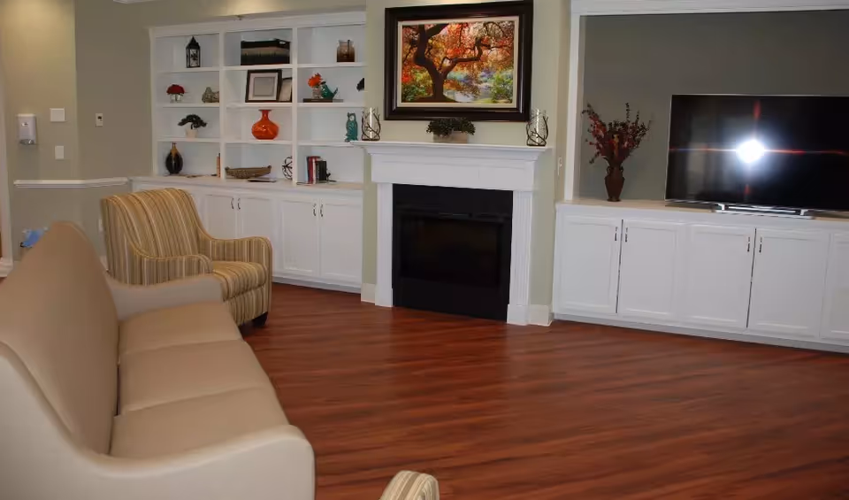 A comfortable common area with a sofa and armchairs facing a fireplace flanked by built-in white shelving and a TV.