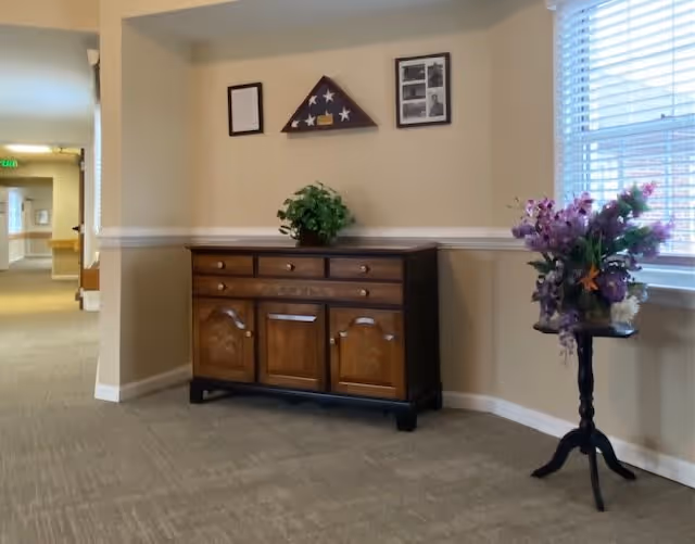Interior hallway area with beige walls and carpeted floor. A wooden cabinet with drawers and doors is against the wall, decorated with a small green plant on top. Above the cabinet, there are framed pictures and a folded American flag in a triangular display case. To the right, a small round table holds a vase with purple flowers, positioned near a window with white blinds. The hallway extends to the left with visible doors and exit signs.
