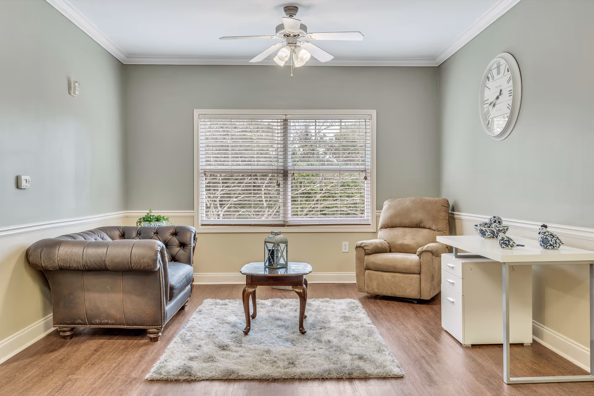 Bright living room with a leather sofa, recliner, coffee table on a rug, and a desk beneath a wall clock.