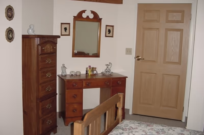 Bedroom with a wooden bed frame, a dresser/vanity topped with small items, a tall chest of drawers, a wall mirror, and a closed wooden door.