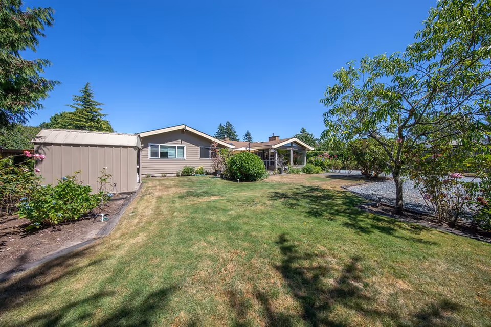 A single-story house with beige siding and a brown roof surrounded by a well-maintained lawn and various trees and shrubs under a clear blue sky.
