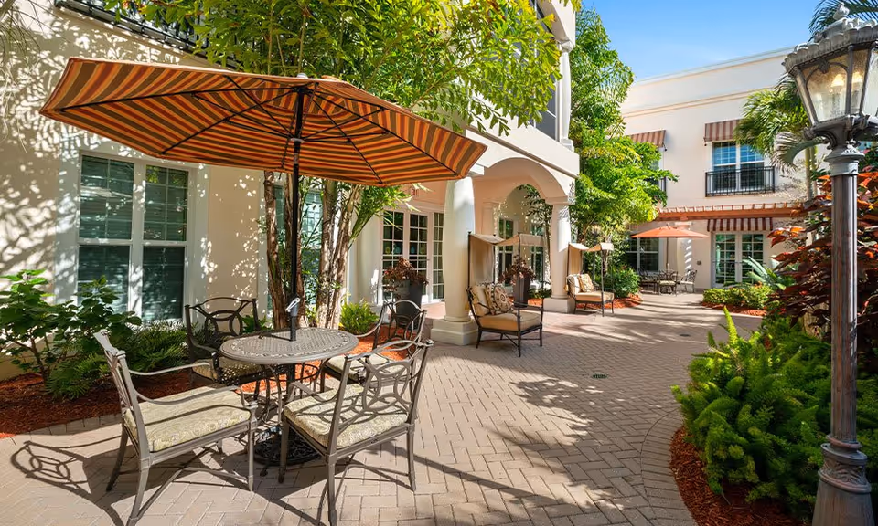 Outdoor courtyard area at Villa at Terracina Grand featuring a round metal table with four cushioned chairs under a large striped umbrella, surrounded by greenery, trees, and shrubs. The courtyard is paved with bricks and has additional seating areas with cushioned chairs and umbrellas near the building with white walls and large windows.