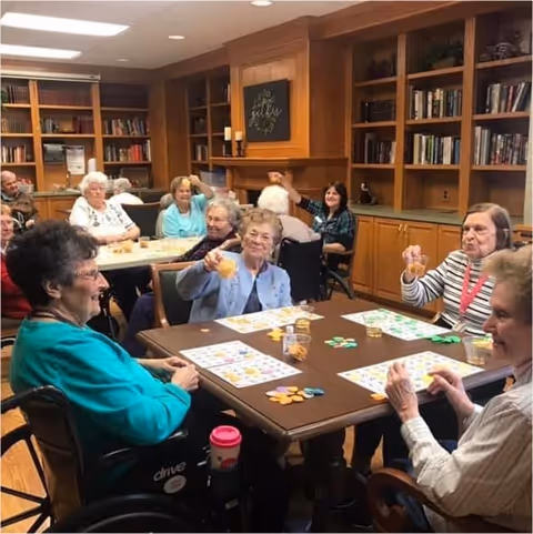 A group of elderly women sitting around tables in a room with wooden bookshelves, playing a game with colorful chips and holding up drinks, smiling and enjoying each other's company.