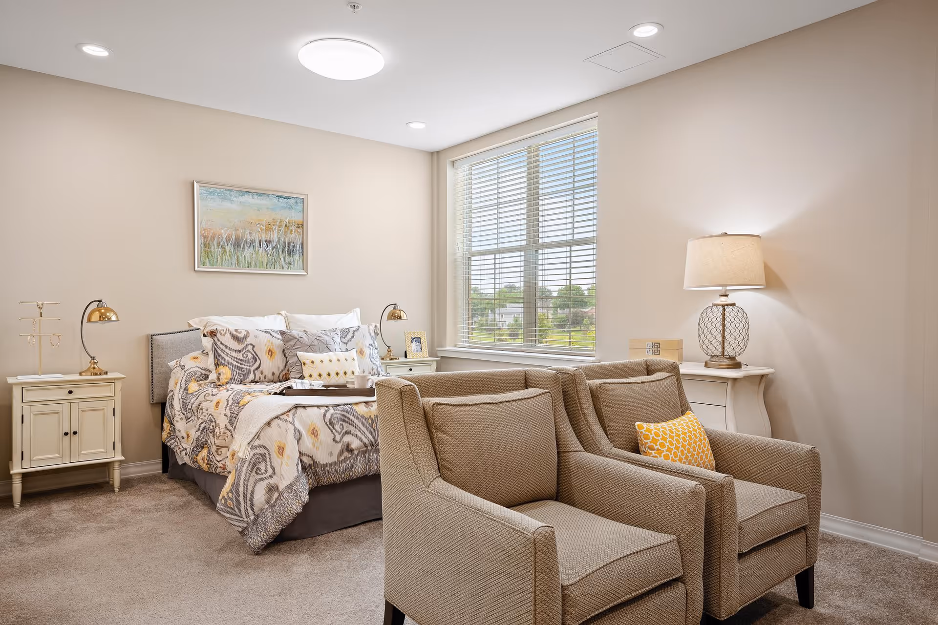 A cozy bedroom with a bed featuring patterned bedding and multiple pillows, flanked by two nightstands with lamps. In front of the bed are two beige armchairs with a yellow patterned pillow on one. A large window with blinds allows natural light into the room, and a framed artwork hangs on the wall above the bed.