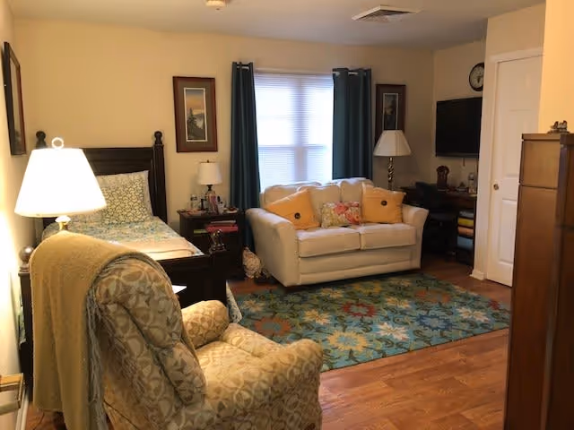 A cozy bedroom with a single bed on the left side, a patterned armchair with a blanket draped over it in the foreground, a white loveseat with colorful pillows in front of a window with dark curtains, a wooden nightstand with a lamp and framed pictures on the wall, a TV mounted on the wall, and a wooden dresser on the right side. The room has wooden flooring and a colorful area rug.