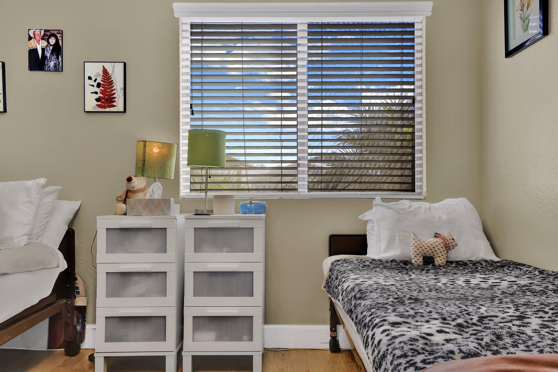 A small bedroom with two single beds separated by two white plastic drawer units. One bed has white pillows and a beige blanket, the other has white pillows and a black and white patterned blanket with a small stuffed dog toy on top. A window with horizontal blinds shows a view of trees and a blue sky with clouds. The walls are light beige with framed pictures hanging on them. A green lamp and a tissue box are on the drawer units.