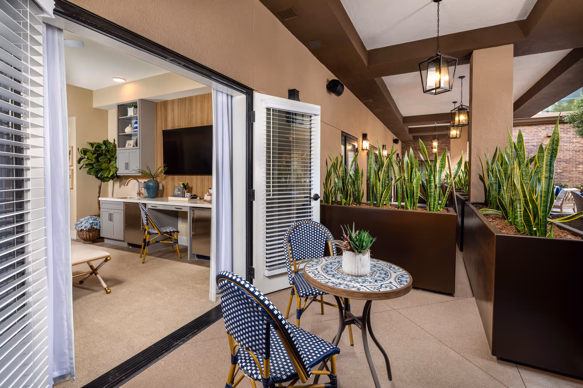 Outdoor patio area with a small round table and two blue and white woven chairs. The patio is decorated with large planters filled with tall green snake plants. Hanging lantern-style lights are suspended from the ceiling. Through an open door, an interior room is visible with a desk, chair, large TV mounted on a wooden accent wall, and a potted plant in the corner.