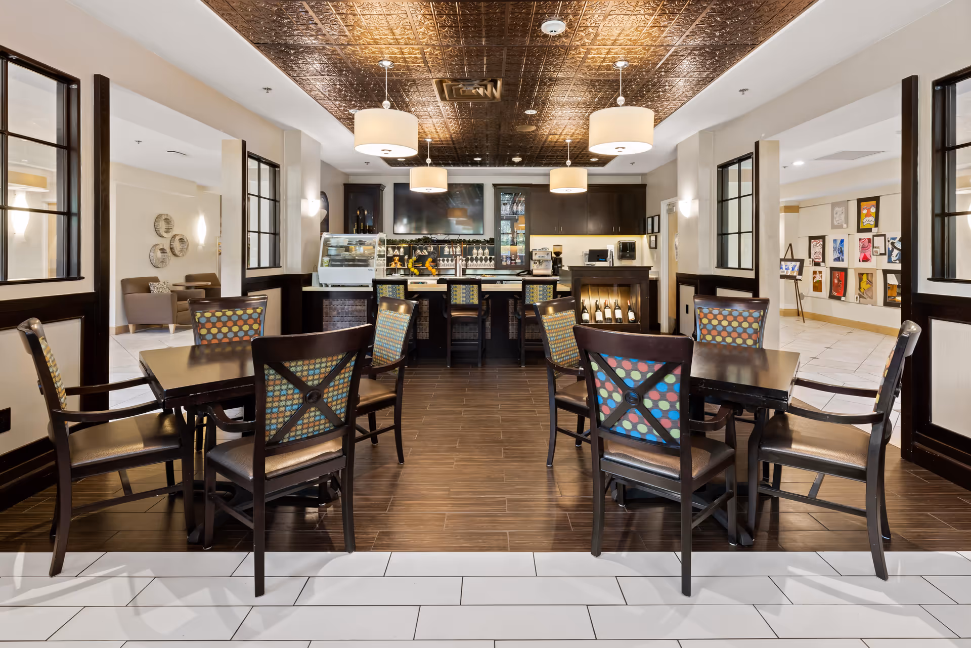 Interior view of a dining area at The Pearl at Boulder Creek featuring dark wooden tables and chairs with colorful patterned backs. The room has a decorative bronze ceiling with hanging pendant lights. In the background, there is a bar counter with stools, a display case, wine bottles, and a large TV mounted on the wall. Adjacent rooms with artwork and seating areas are visible through open doorways.