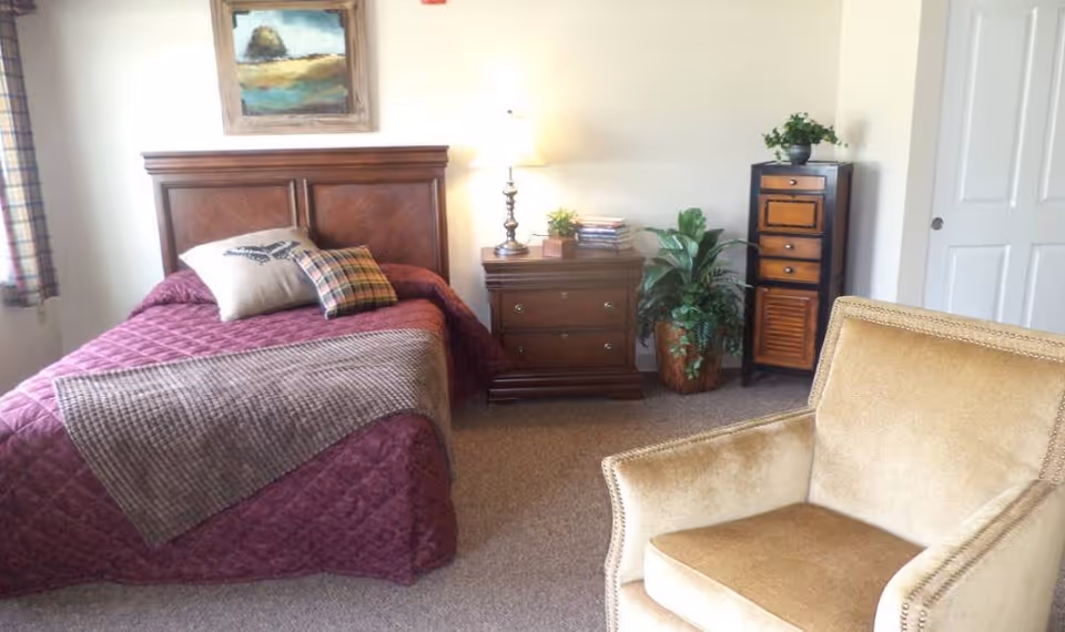 A cozy bedroom with a single bed covered in a maroon quilt and two pillows, one beige and one plaid. Next to the bed is a wooden nightstand with a lamp, a small plant, and some books. There is a tall plant in a pot on the floor beside a small wooden cabinet with drawers. A beige upholstered armchair is positioned in the foreground. A framed painting hangs on the wall above the bed, and a window with plaid curtains is partially visible on the left.