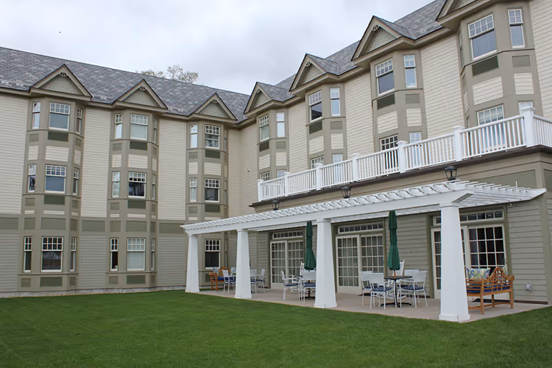 Exterior view of a multi-story assisted living facility with beige and green siding, multiple windows, and a covered patio area with tables, chairs, and umbrellas on a well-maintained lawn.