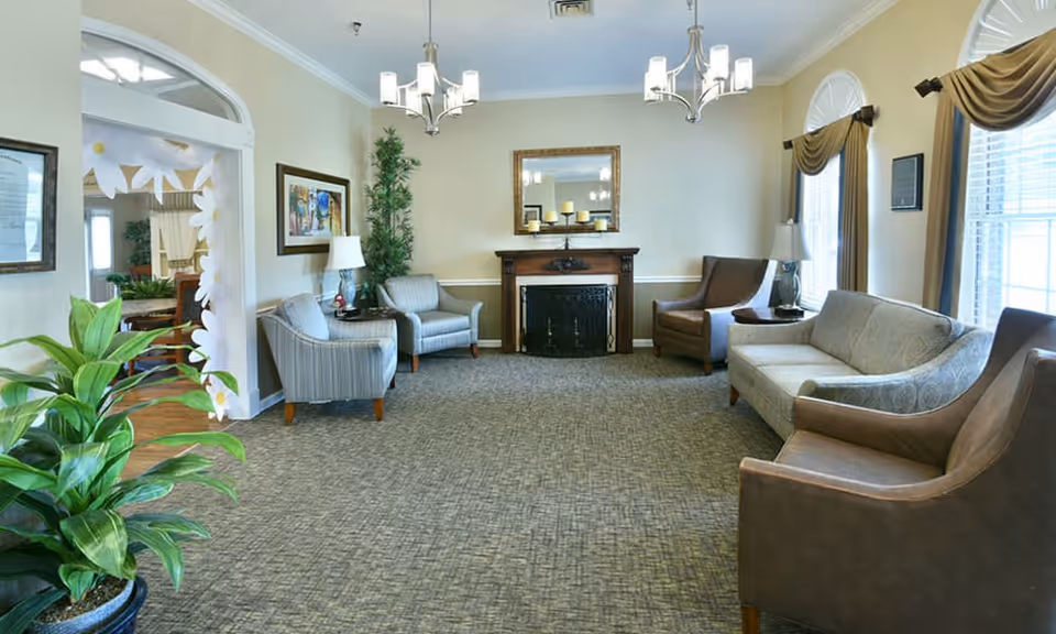 A well-lit senior living facility common area with several upholstered chairs and a sofa arranged around a fireplace with a mirror above it. There are two table lamps on side tables, large windows with drapes, and green plants adding to the decor. The room has a carpeted floor and light-colored walls.