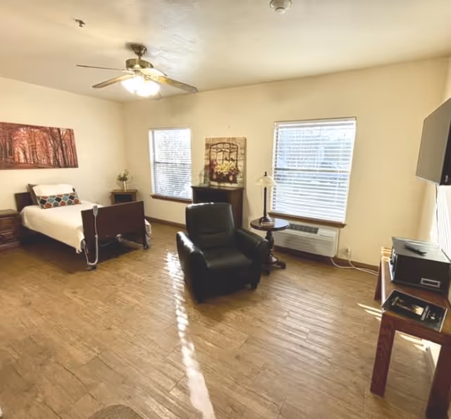 Bright, spacious bedroom with a single bed, leather armchair, side tables, windows with blinds, and a wall-mounted TV.