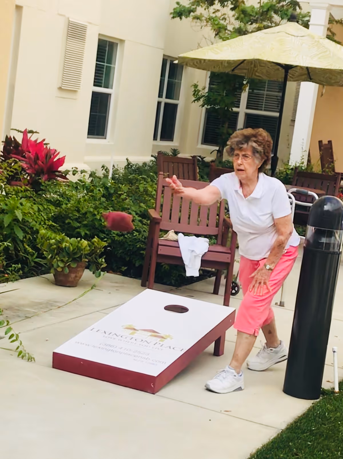 An elderly woman wearing a white shirt and pink pants is playing cornhole outdoors on a concrete patio. She is in the motion of tossing a bean bag towards a cornhole board that has the Lexington Place logo and contact information on it. There are wooden chairs, green plants, and a large umbrella in the background near a building with windows.
