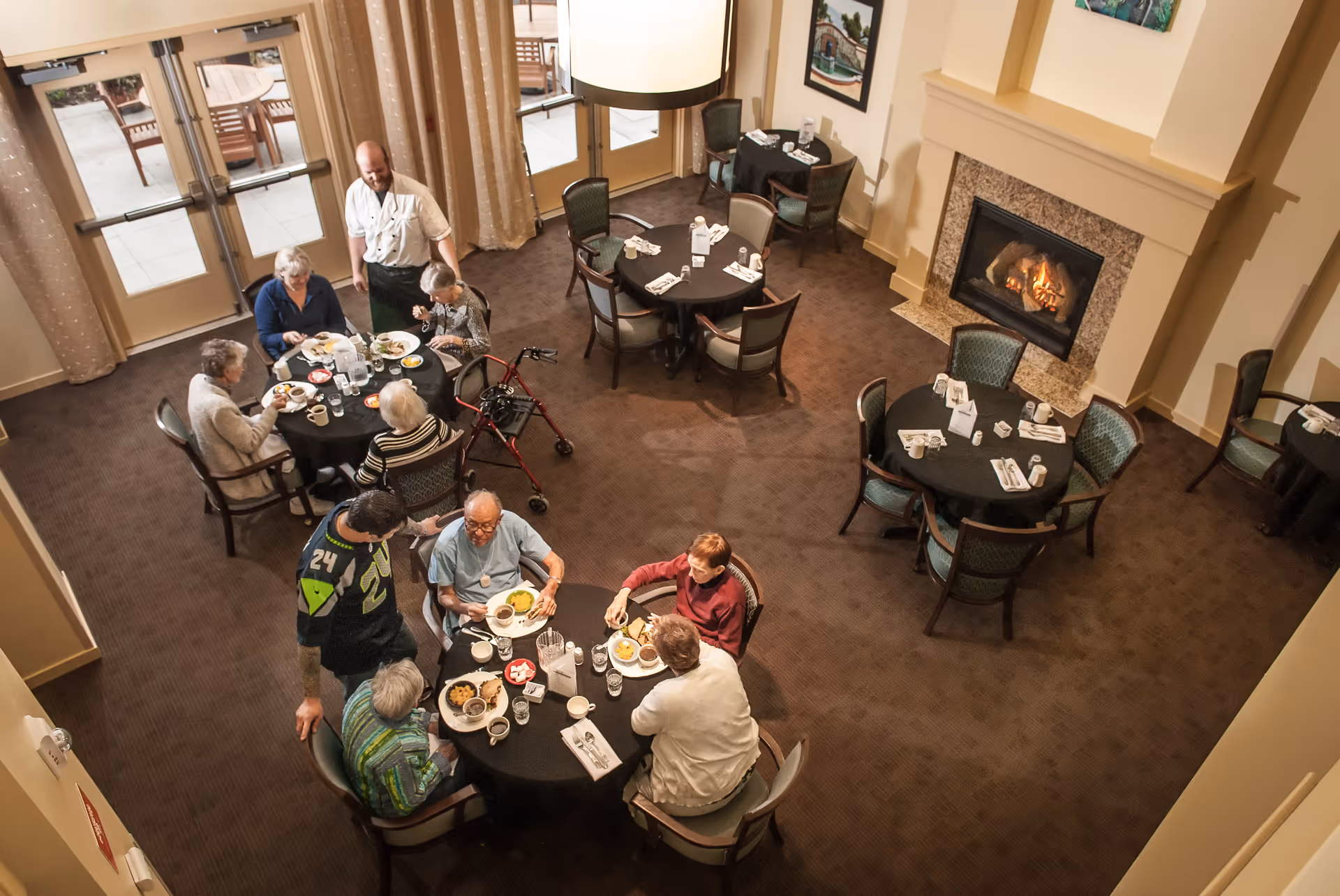 Overhead view of a senior dining room with several elderly people seated at round tables enjoying a meal. A staff member is standing and interacting with one of the tables. The room features a lit fireplace, carpeted floor, and large windows with curtains leading to an outdoor patio area.