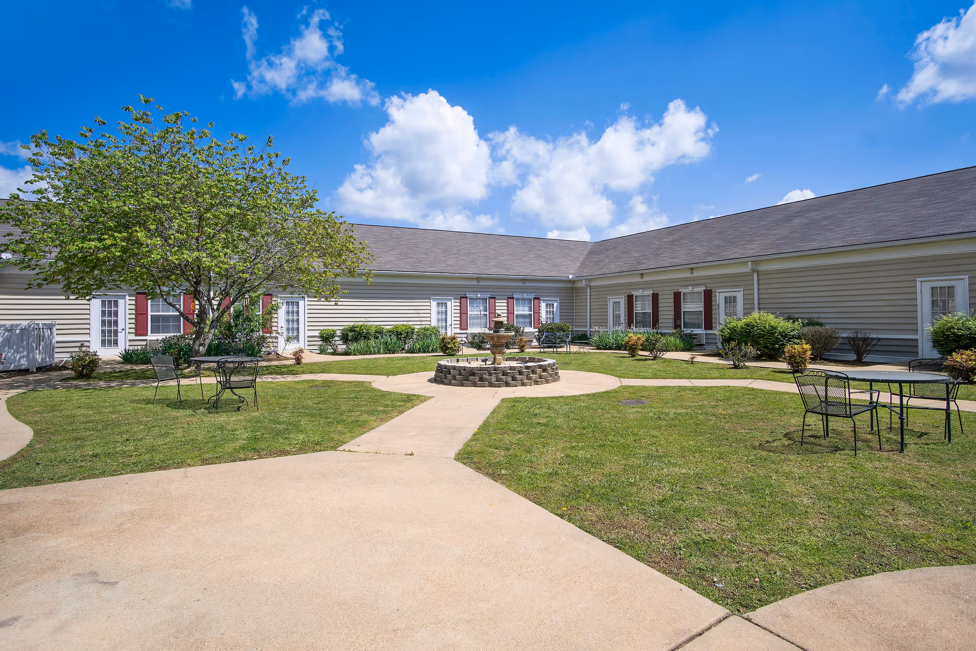 Outdoor courtyard area at HarborChase of Jasper featuring a circular stone fountain in the center, surrounded by green grass and paved walkways. There are metal tables and chairs placed around the courtyard, with a tree providing some shade. The building exterior has beige siding with white doors and windows accented by red shutters under a clear blue sky with some clouds.
