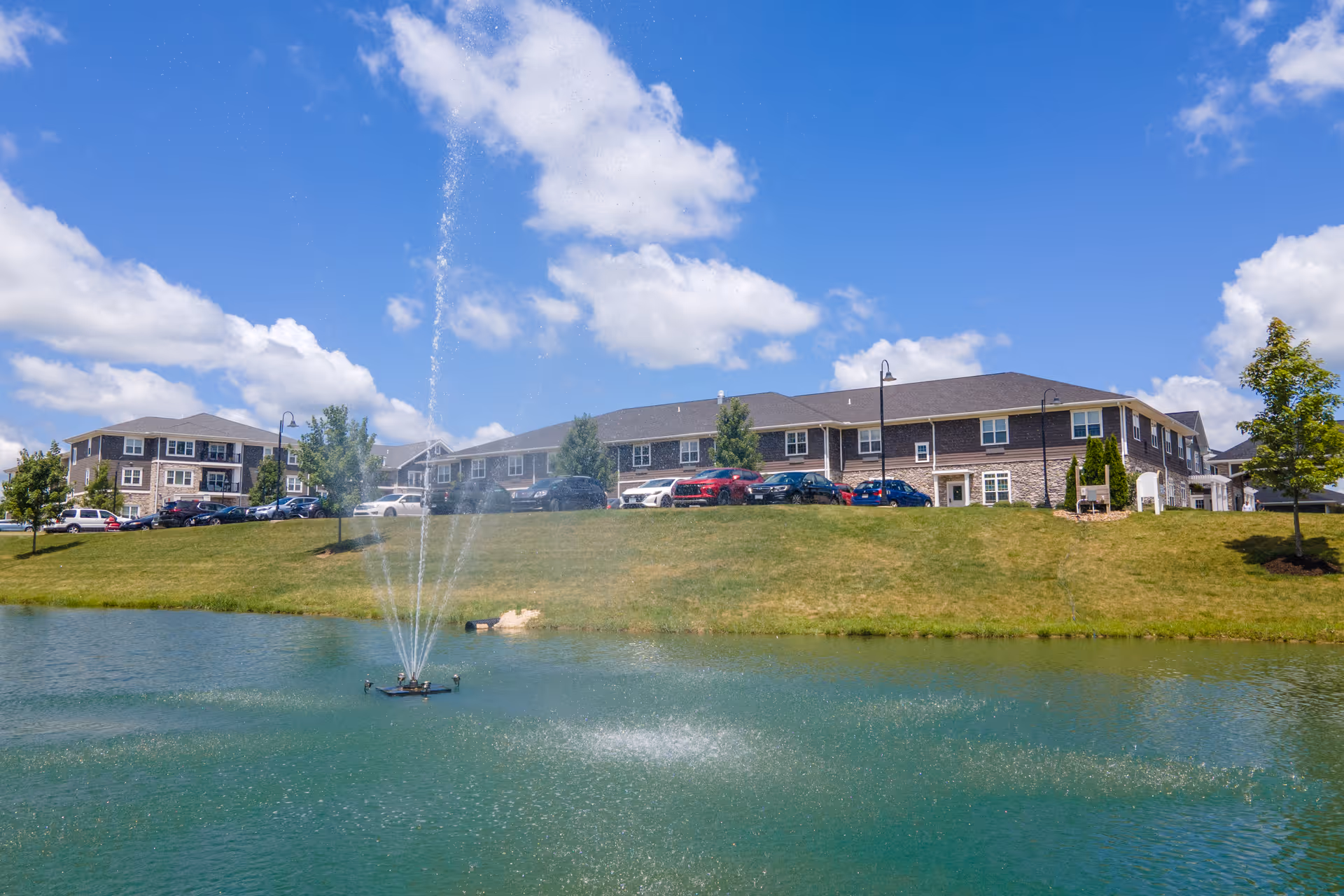 Low-rise residential building behind a grassy slope with a pond and a central fountain under a blue sky.