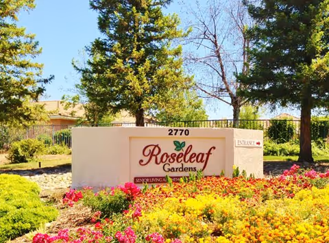 Outdoor view of the entrance sign for Roseleaf Gardens senior living facility surrounded by colorful flowers and greenery with trees and a clear blue sky in the background.