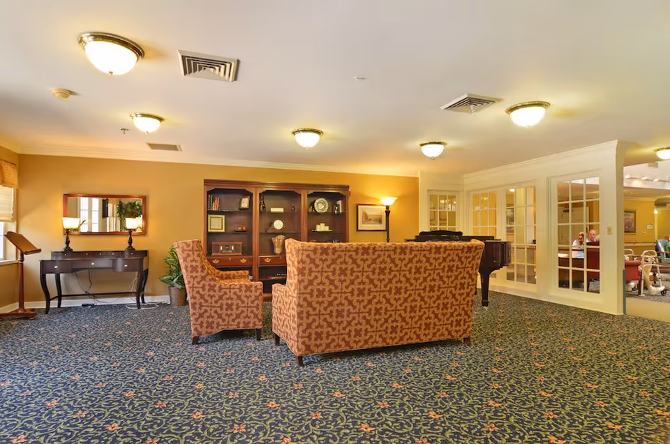 A cozy living room area in a senior living facility with patterned carpet, two upholstered armchairs and a matching sofa facing a wooden bookshelf filled with decorative items. There is a wooden console table with two lamps and a mirror on the left wall, a floor lamp in the corner, and a grand piano near a set of glass-paneled doors. The room is warmly lit with ceiling lights and has a calm, inviting atmosphere.