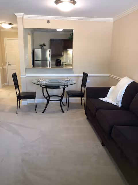 Interior view of a senior living facility showing a small dining area with a round glass table set for two and two black chairs. To the right, there is a dark brown sofa with a white throw blanket. In the background, a kitchen area with a stainless steel refrigerator, wooden cabinets, and a coffee maker is visible through a pass-through window. The walls are beige, and the floor is carpeted.