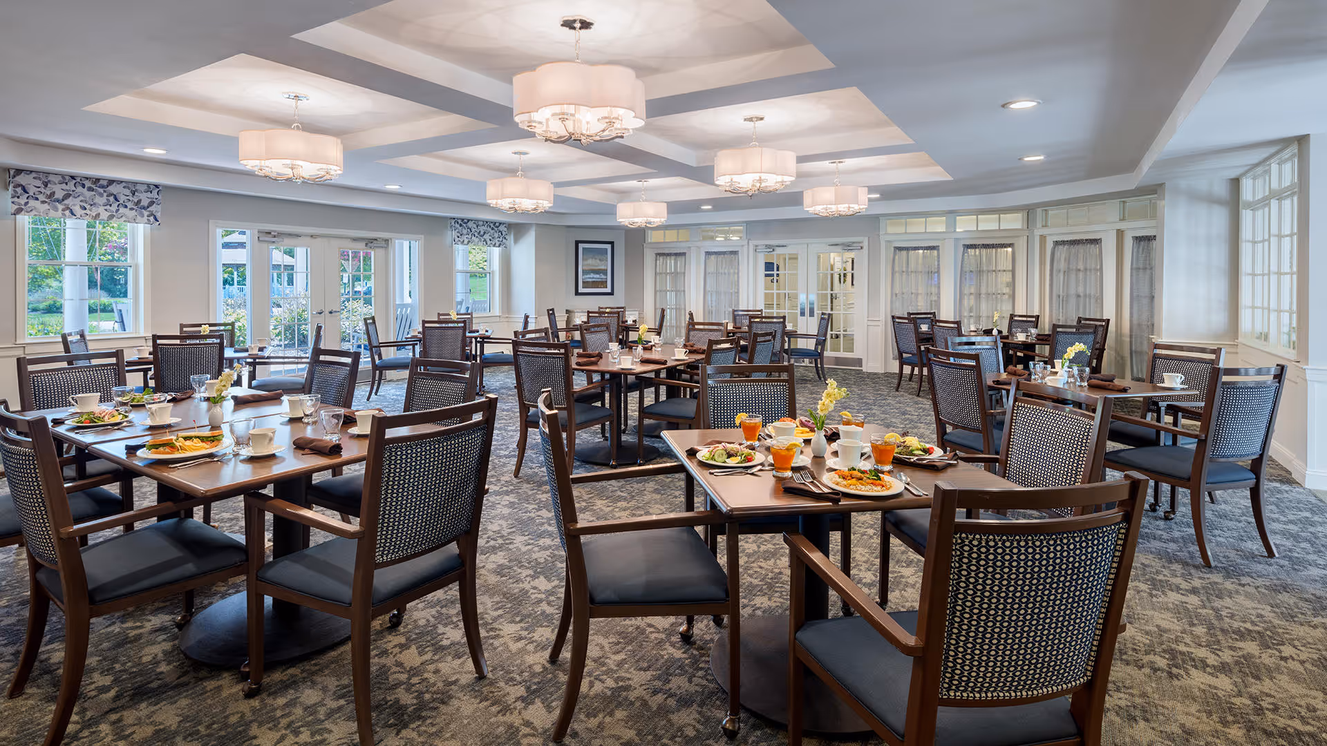 A spacious dining room with multiple wooden tables and chairs arranged neatly. Each table is set with plates of food, glasses of orange juice, cups, and napkins. The room has large windows with floral valances, allowing natural light to brighten the space. The ceiling features recessed lighting and decorative light fixtures. The carpet has a patterned design, and the walls are painted in light colors with white trim.