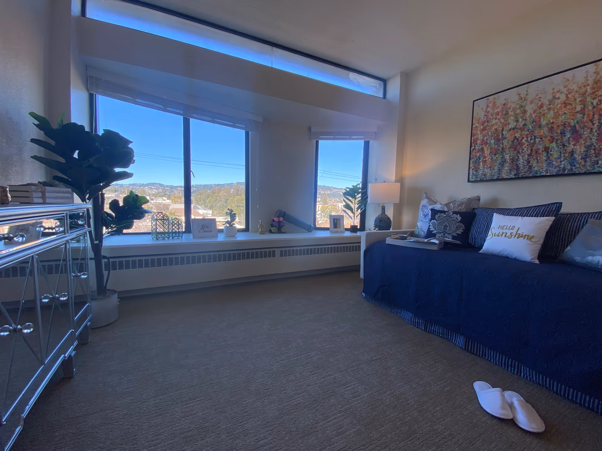 A cozy room with a large window showing a scenic outdoor view. The room features a blue daybed with decorative pillows, including one that says 'Hello Sunshine.' There is a lamp on a side table next to the bed, a mirrored dresser with books and a plant on top, and a pair of white slippers on the carpeted floor. A colorful floral painting hangs on the wall above the bed.