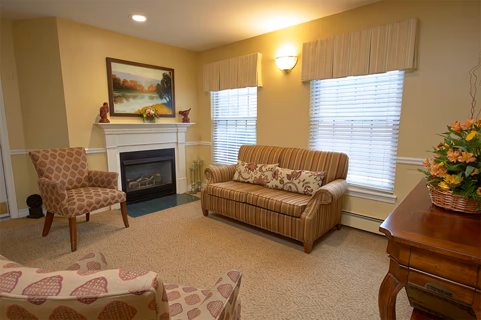 A cozy living room with a striped sofa adorned with floral cushions, two patterned armchairs, a fireplace with decorative items on the mantel, two windows with blinds and valances, a wall-mounted light fixture, and a wooden table with a basket of flowers.
