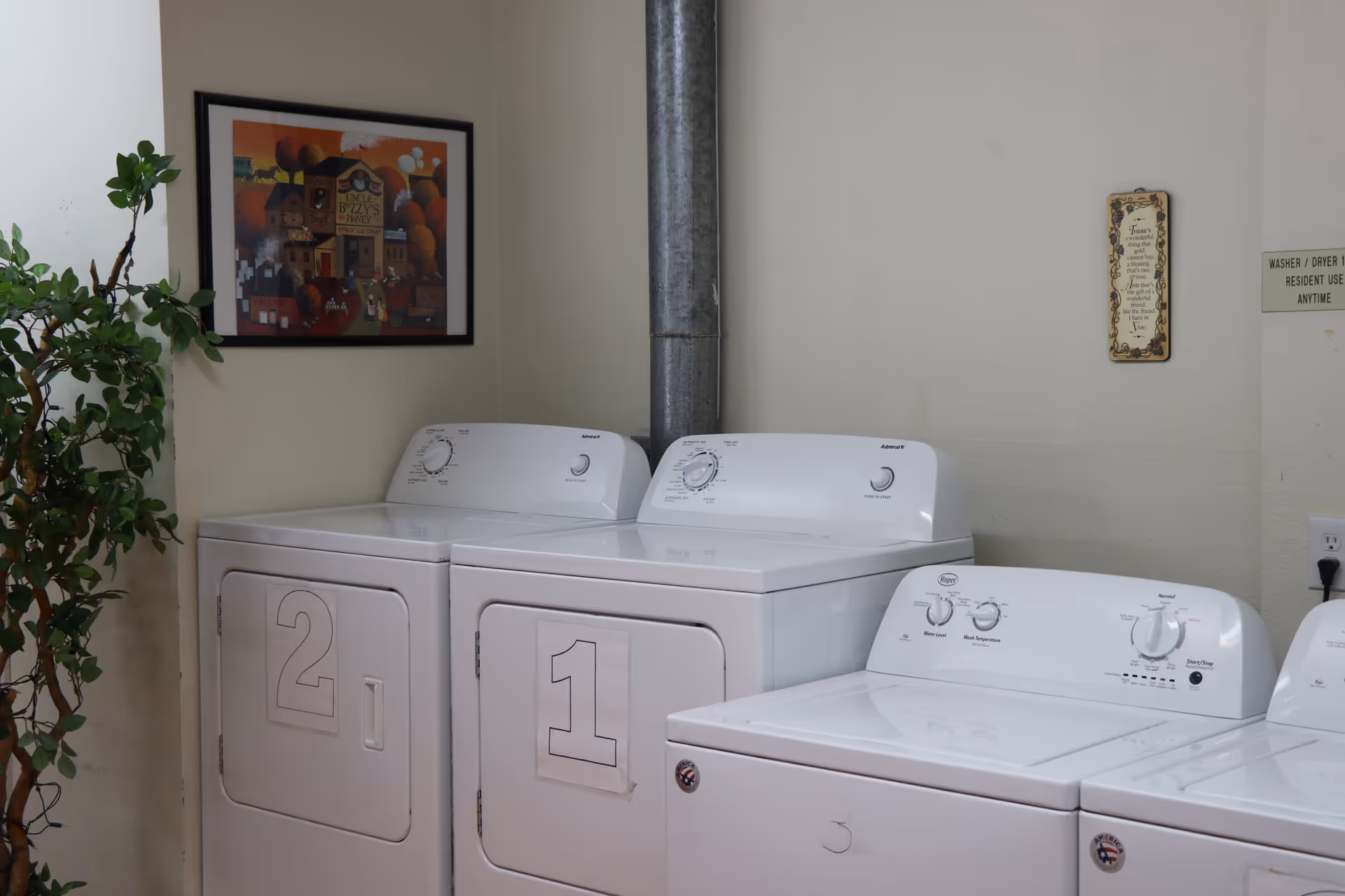 Laundry room with two white dryers labeled 1 and 2, and two white washing machines. A green potted plant is on the left side, and a framed picture hangs on the wall above the dryers. There are two signs on the wall, one with a decorative quote and another indicating washer/dryer use instructions.