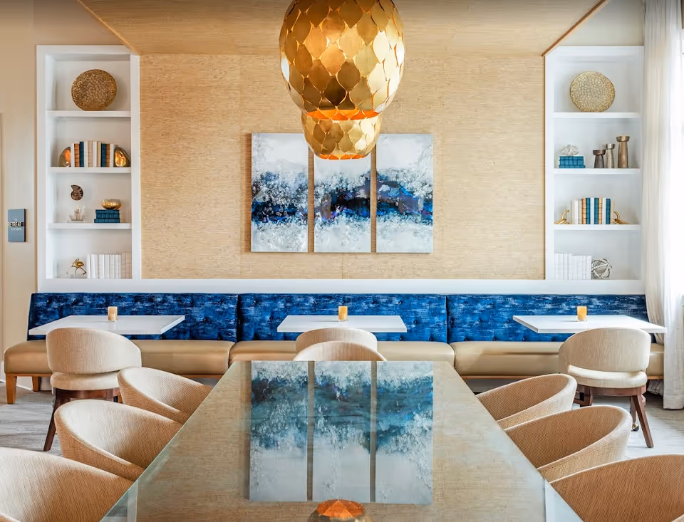 Bright dining area with a glass-top table, upholstered chairs, blue banquette seating, and gold pendant lights.
