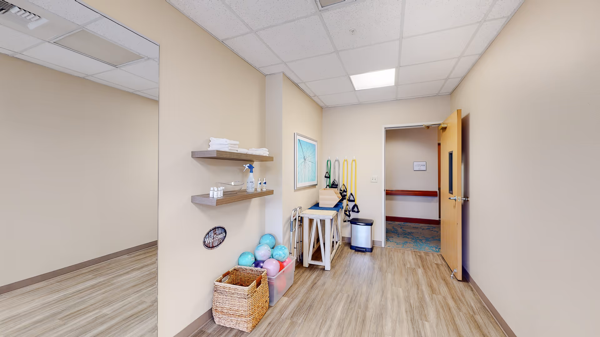 A small exercise or therapy room with wooden flooring and beige walls. The room contains shelves with towels and cleaning supplies, a basket with colorful exercise balls, resistance bands hanging on the wall, a small table with equipment, and an open door leading to a hallway.