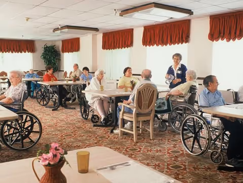 A group of elderly people, many in wheelchairs, sitting around tables in a well-lit dining room with large windows covered by red valances. A caregiver is standing and interacting with some of the residents. The room has patterned carpet and simple decor.