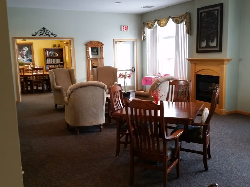 A cozy common area with a wooden table and four chairs in the foreground, beige upholstered armchairs arranged near a fireplace with a wooden mantel, a tall mirror against the wall, and a window with white curtains and a gold valance. In the background, there is an adjoining room with bookshelves and a dining table with chairs.