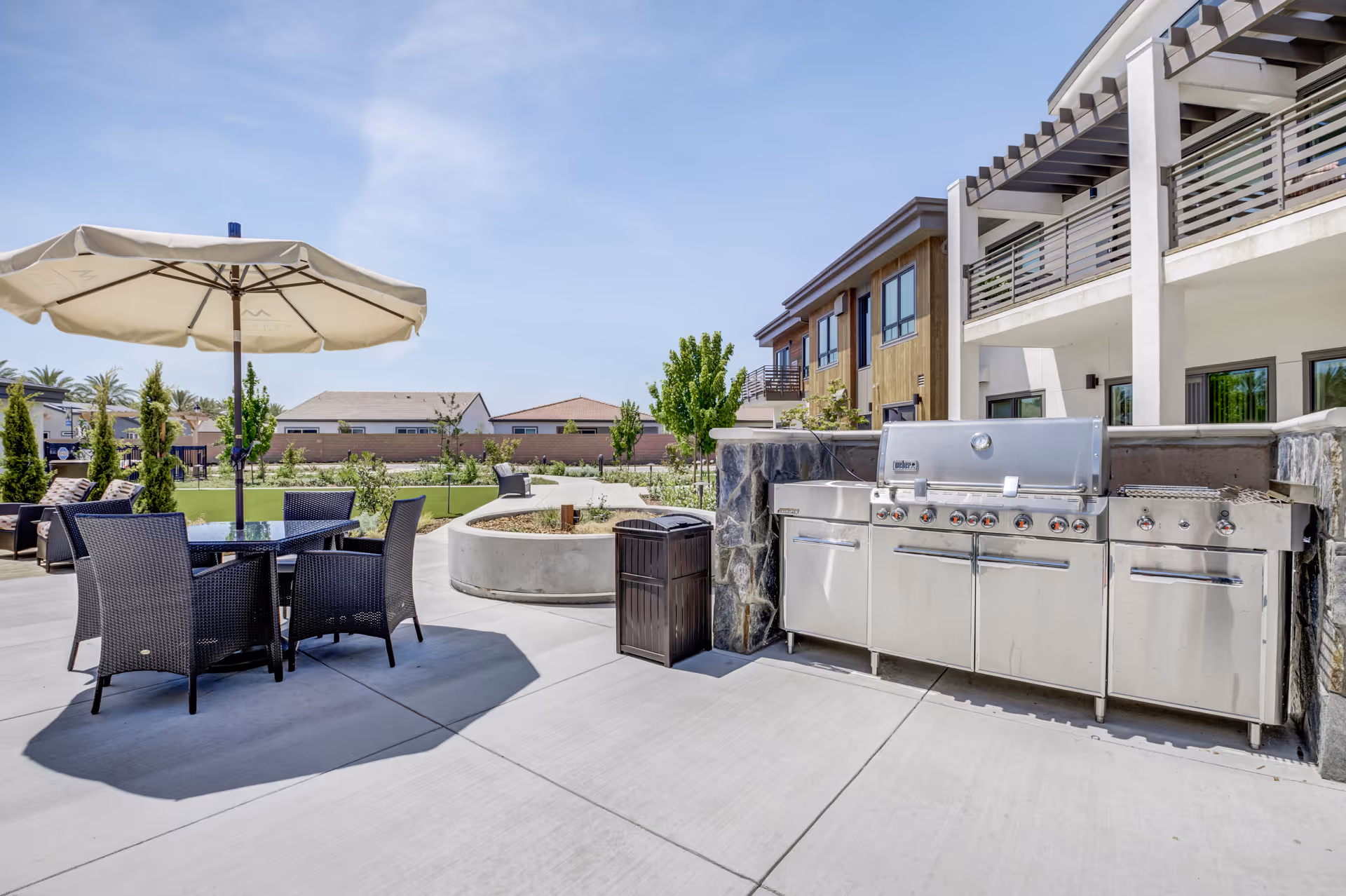 Outdoor patio area with a large stainless steel barbecue grill, a round glass table with four wicker chairs, and a large beige umbrella providing shade. In the background, there are green lawns, small trees, and a multi-story building with balconies under a clear blue sky.