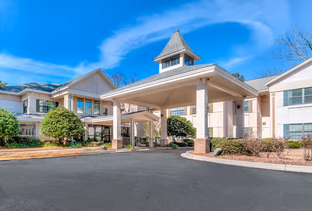 Exterior view of Oaks at Habersham facility showing a covered entrance with a peaked roof, surrounded by a two-story building with multiple windows, shrubs, and trees under a blue sky with some clouds.