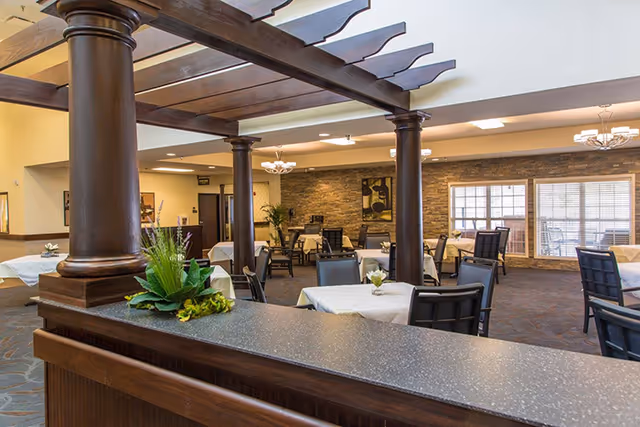 Interior view of a dining area in a senior living facility with tables covered in white tablecloths, black chairs, decorative plants, wooden columns, and a stone accent wall with framed artwork.
