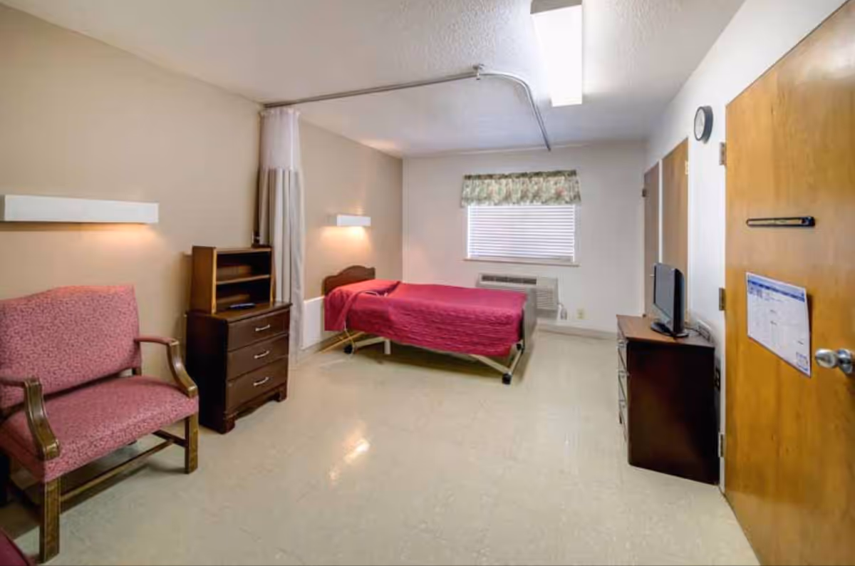 Hospital-style bedroom with single bed, chair, dresser, TV, and window.
