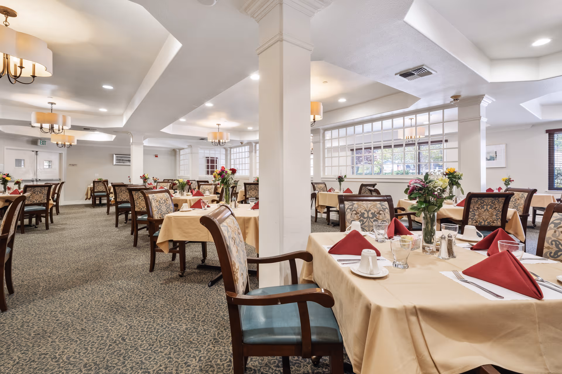 A spacious dining room with multiple tables covered in beige tablecloths, each set with red folded napkins, cups, glasses, and silverware. The chairs have patterned upholstery and wooden armrests. The room is well-lit with ceiling lights and chandeliers, and there are flower arrangements on each table. Large windows and white pillars are visible in the background.