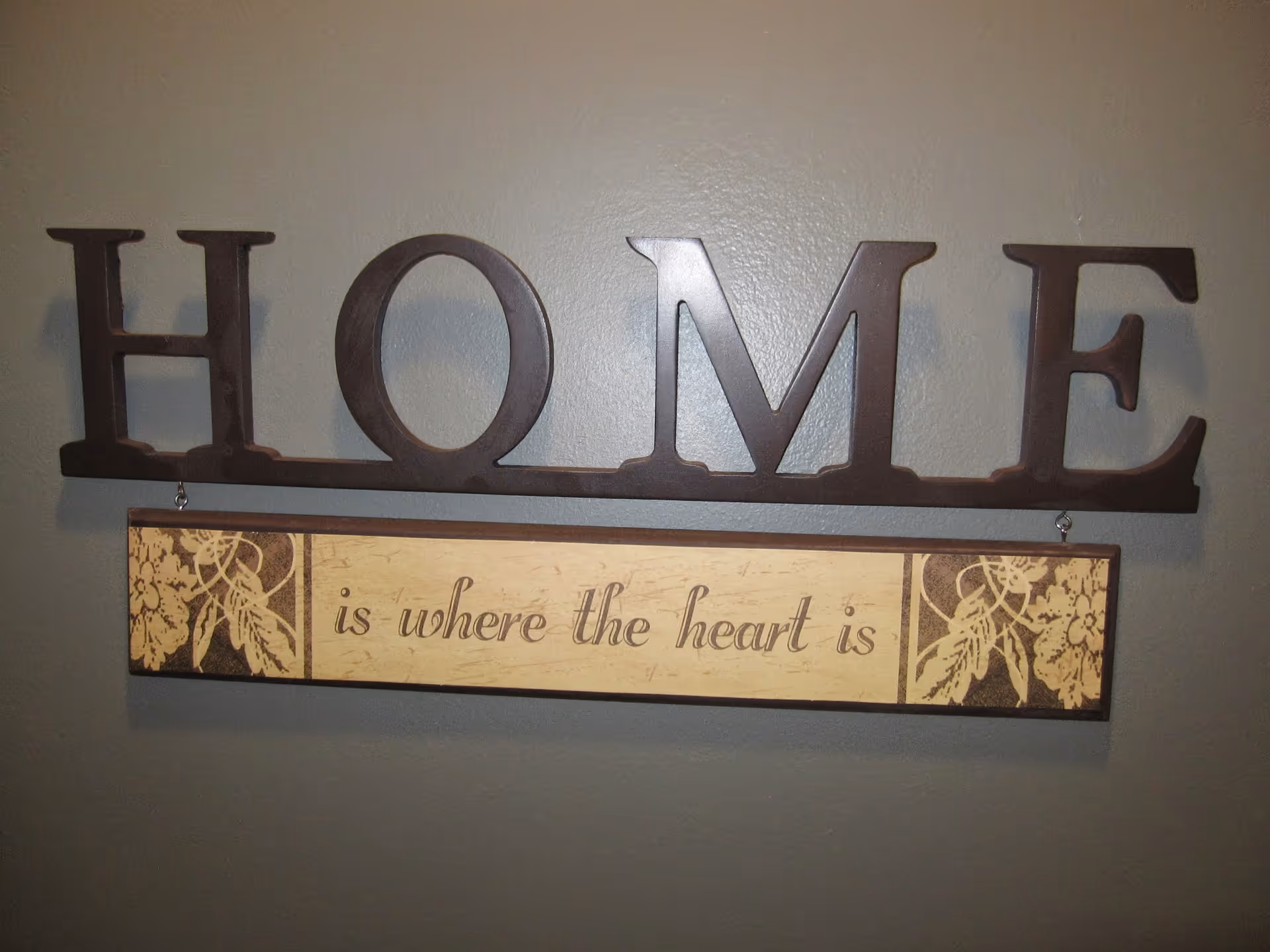 A decorative wall sign with the word 'HOME' in large brown letters above a smaller plaque that reads 'is where the heart is' with floral designs on either side, mounted on a gray wall.