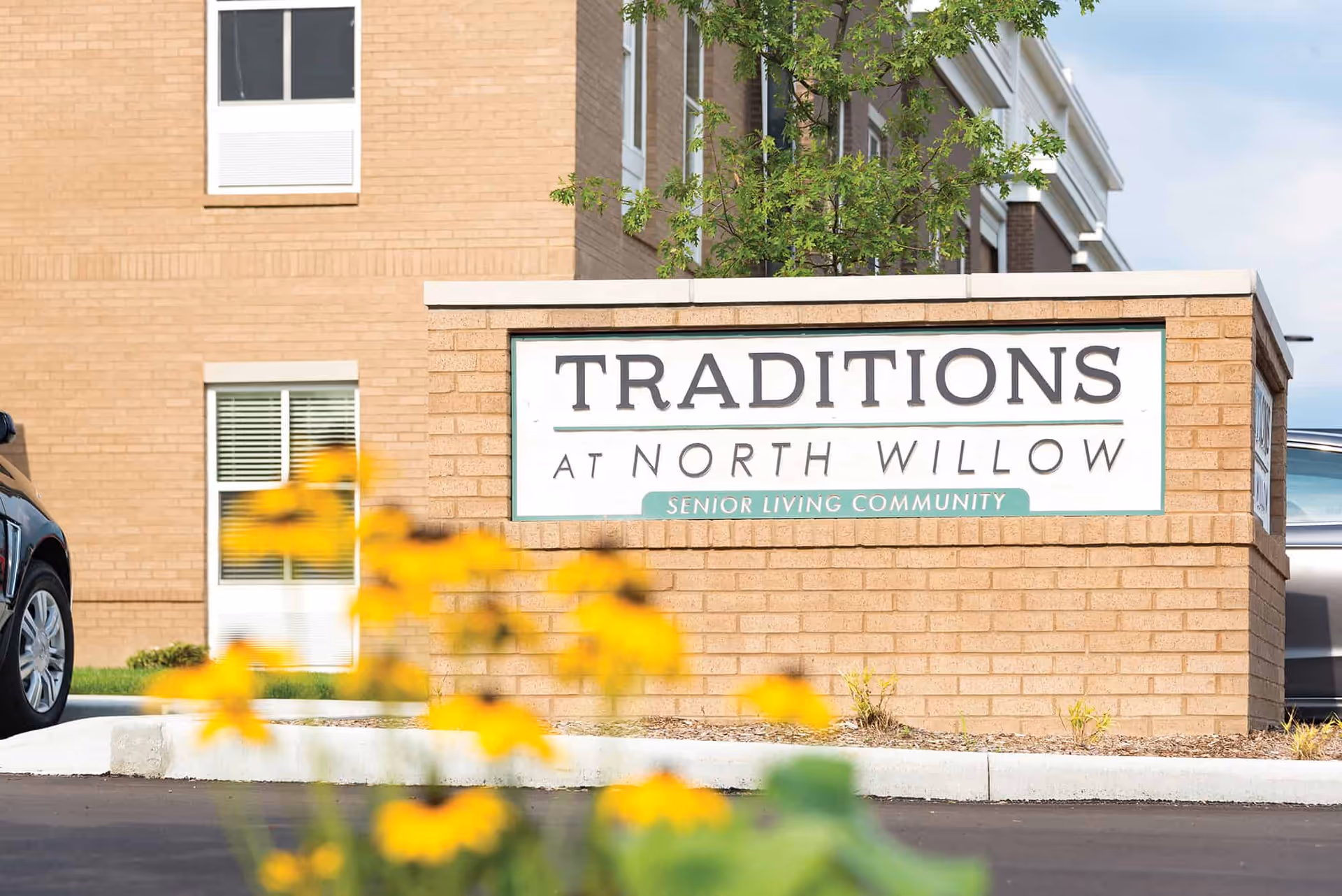 A brick sign outside a building that reads 'Traditions at North Willow Senior Living Community' with yellow flowers in the foreground and part of a car visible on the left side.