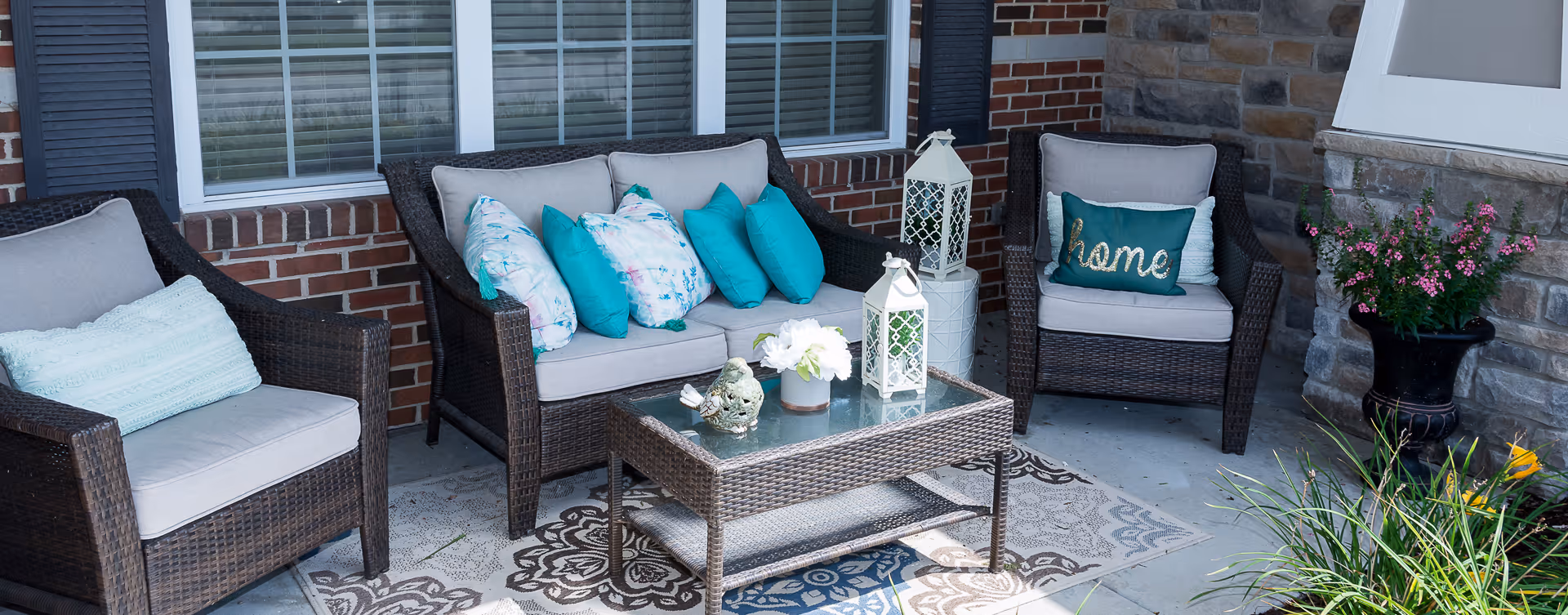 Outdoor patio area with wicker furniture including a loveseat and two armchairs with light gray cushions and teal and floral throw pillows. A glass-top wicker coffee table holds decorative items including a small bird figurine, a white flower arrangement, and two white lanterns. The patio is adjacent to a brick and stone wall with a window and has a patterned outdoor rug on the floor. There is a black planter with pink flowers and some green plants nearby.