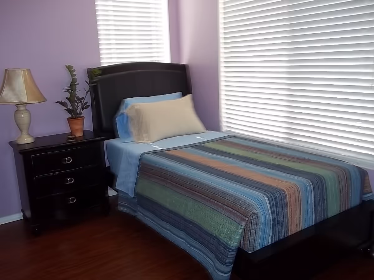 A neatly made single bed with a striped bedspread next to a dark nightstand, lamp, and potted plant in a room with purple walls and white blinds.