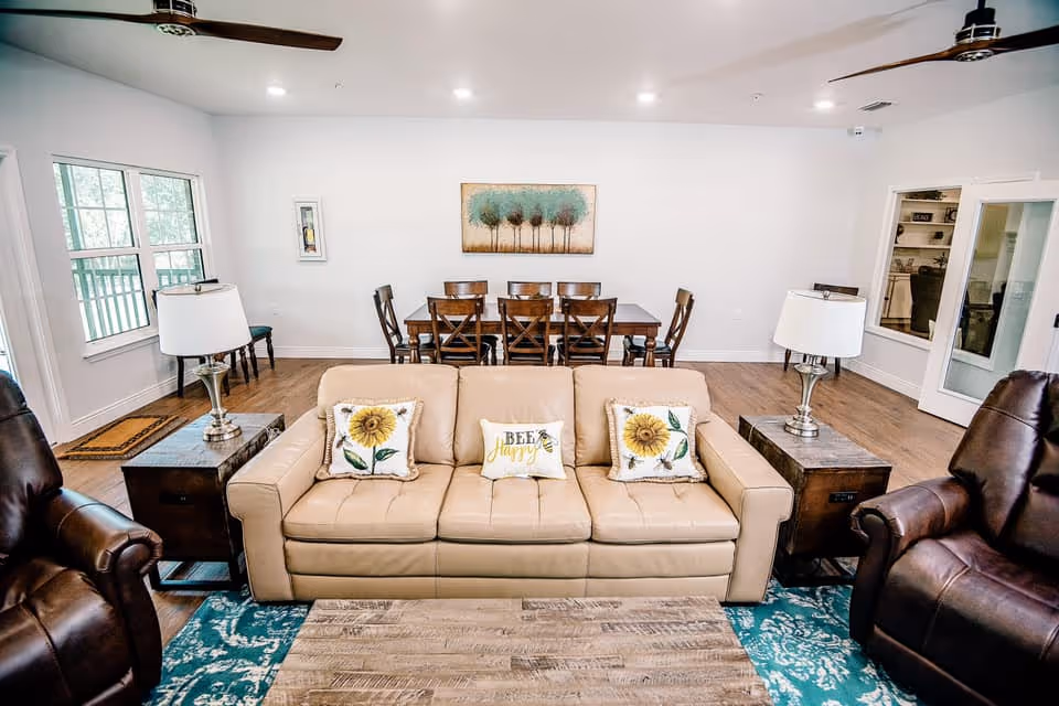 Bright communal living room with a beige sofa and sunflower pillows, recliners, coffee table, and a dining table in the background.