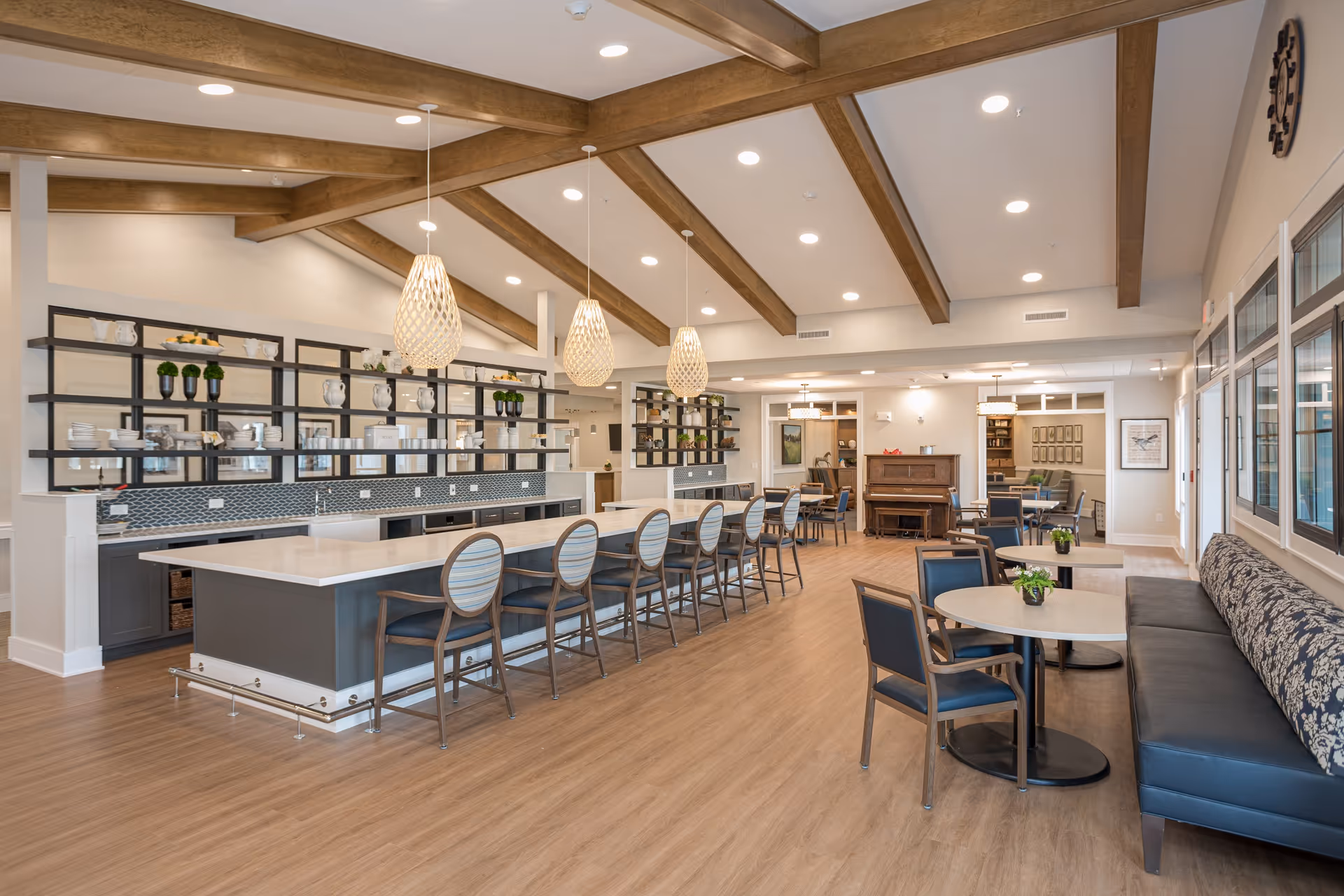 Spacious and well-lit common area in a senior living facility featuring a long kitchen island with bar stools, pendant lights hanging from a ceiling with wooden beams, round tables with chairs, a cushioned bench along the window, and a piano in the background.
