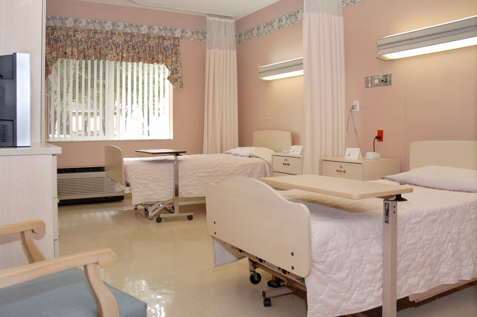 Two-bed medical-style patient room with pastel walls, two hospital beds, overbed tables, nightstands and a window with vertical blinds.