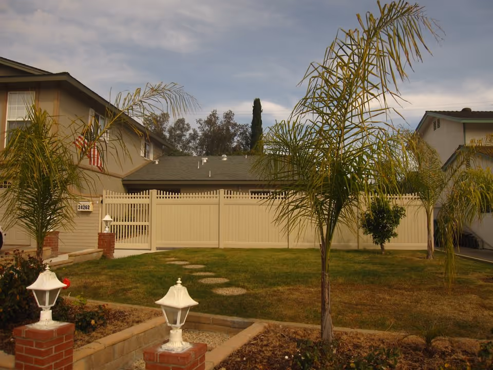 Front yard of a residential building with a green lawn, small palm trees, a beige fence, and a two-story house with an American flag on the left side.